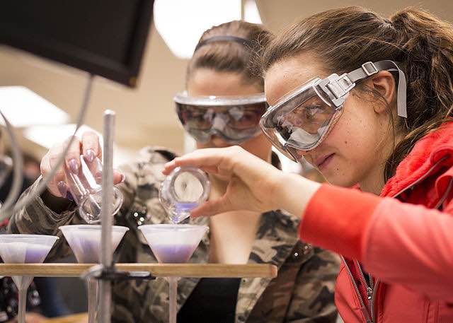 two students with safety goggles pouring liquid into flasks
